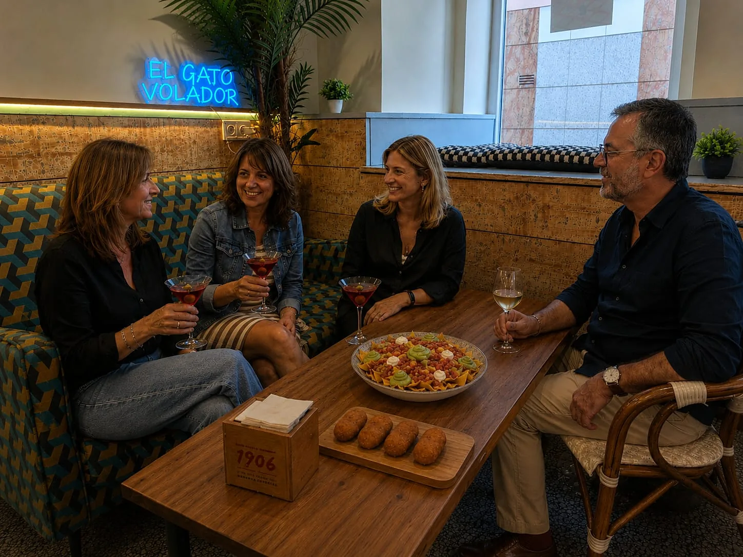 Mesa con cócteles servidos en copa, una copa de vino blanco, nachos y croquetas bajo el neón del bar